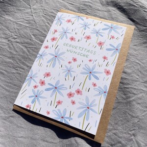 May include: A greeting card with a floral design featuring blue and pink flowers, with the German text "GEBURTSTAGS W&Uuml;NSCHE" in green. The card is set inside a brown envelope, perfect for sending birthday wishes.
