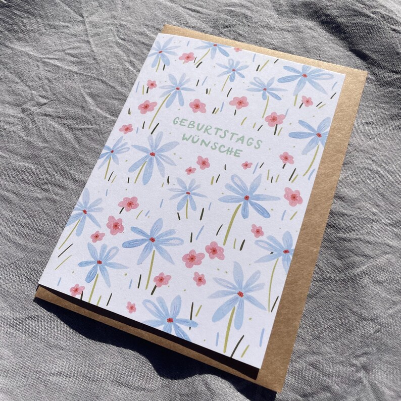 May include: A greeting card with a floral design featuring blue and pink flowers, with the German text "GEBURTSTAGS W&Uuml;NSCHE" in green. The card is set inside a brown envelope, perfect for sending birthday wishes.
