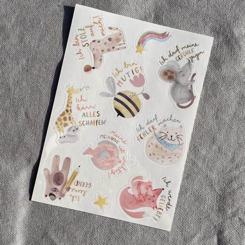K&ouml;nnte beinhalten: Ein Stickerbogen mit Aquarellillustrationen von Tieren und inspirierenden S&auml;tzen auf Deutsch. Designs umfassen eine Giraffe, eine Biene, eine Katze, einen Fuchs und eine Maus, mit Texten wie "Ich bin mutig" und "Ich werde geliebt!"