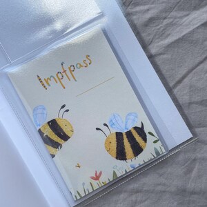 May include: A protective clear plastic sleeve containing a white vaccination record booklet titled Impfpass. The cover features two illustrated yellow and black bumblebees with light blue wings, surrounded by small colorful flowers and green grass.