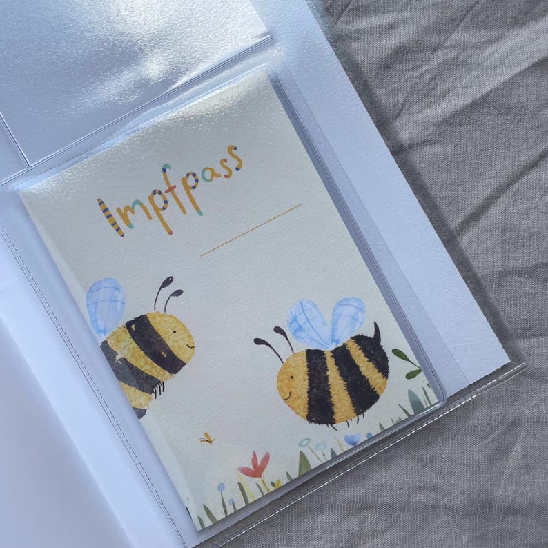 May include: A protective clear plastic sleeve containing a white vaccination record booklet titled Impfpass. The cover features two illustrated yellow and black bumblebees with light blue wings, surrounded by small colorful flowers and green grass.