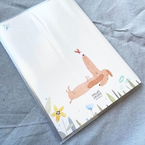 May include: A stack of white paper sheets featuring a watercolor illustration of a dachshund dog reaching for a butterfly. The dog is light brown with darker brown ears. The illustration includes flowers and greenery.