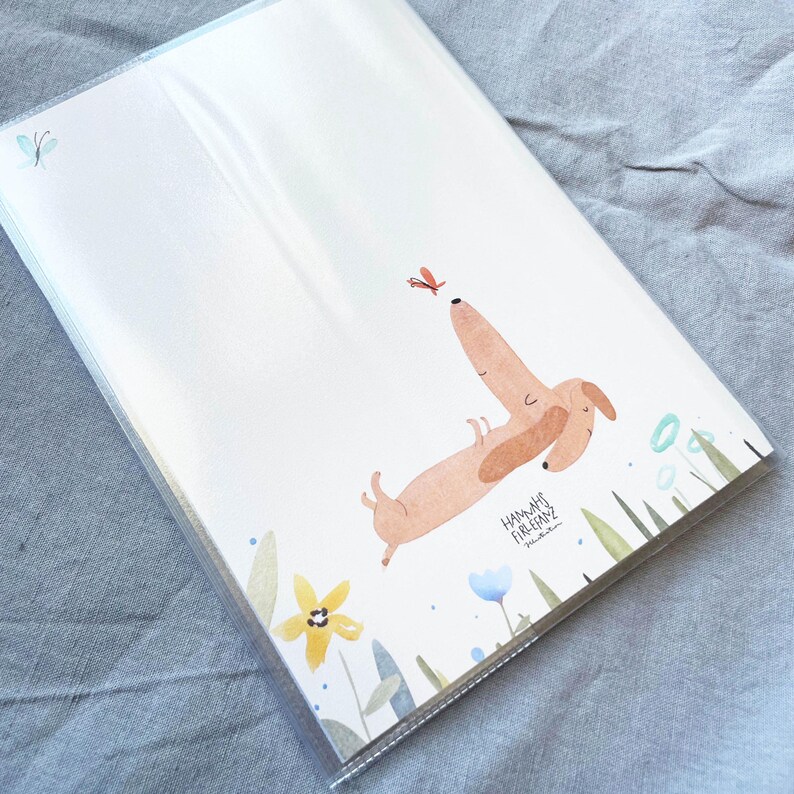 May include: A stack of white paper sheets featuring a watercolor illustration of a dachshund dog reaching for a butterfly. The dog is light brown with darker brown ears. The illustration includes flowers and greenery.