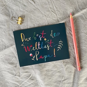 May include: A dark blue card with the text "Du bist der weltbeste Papa!" in colorful lettering. The card is decorated with small flowers, bees, and leaves.