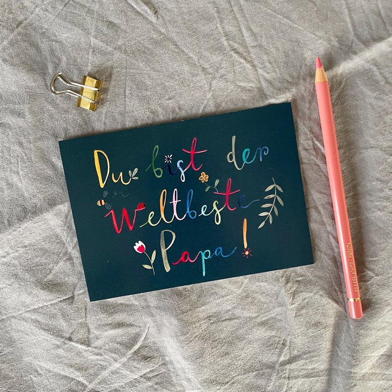 May include: A dark blue card with the text "Du bist der weltbeste Papa!" in colorful lettering. The card is decorated with small flowers, bees, and leaves.