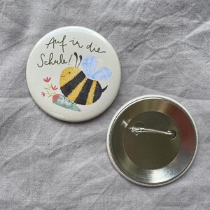 May include: A round pinback button featuring a cream-colored front with a black and yellow bee illustration, a small floral sprig, and the text "Auf in die Schule!" in black script. The back of the button is metallic silver with a pin mechanism.