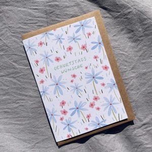 May include: A greeting card with a floral design featuring blue and pink flowers on a white background. The card is paired with a brown envelope. The text "GEBURTSTAGS W&Uuml;NSCHE" is printed in green on the card.