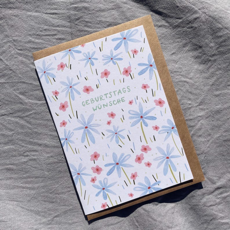 May include: A greeting card with a floral design featuring blue and pink flowers on a white background. The card is paired with a brown envelope. The text "GEBURTSTAGS W&Uuml;NSCHE" is printed in green on the card.