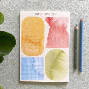 Peut inclure: Un bloc-notes avec les mots "NOTES + DOODLES" imprimés en haut. Le bloc-notes comporte quatre sections différentes pour les notes et les gribouillis. Les sections sont un rectangle jaune avec des lignes, une forme irrégulière rouge avec des points blancs, une grille bleue et un ovale vert. Deux crayons de couleur sont posés sur une surface grise à côté du bloc-notes.