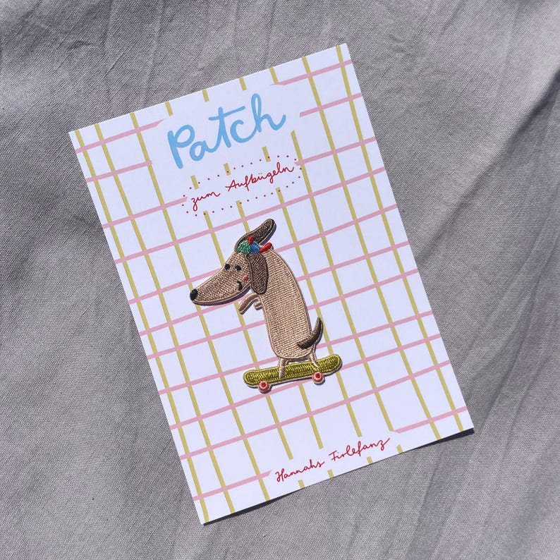 May include: Embroidered patch featuring a dachshund dog on a skateboard. The patch is on a card with the word "Patch" in blue, and text in German. The card has a pink and yellow grid pattern, with the name "Hannahs Friefanz" in red.