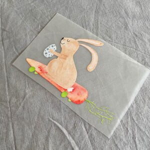 May include: A brown bunny rabbit with a white egg in its paws is riding a carrot-shaped skateboard with green wheels. The carrot has green leaves.