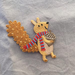 May include: Embroidered patch featuring a cartoon squirrel holding an acorn. The squirrel is tan with a red, white, and blue striped scarf. The patch has a detailed, textured design and is set against a light blue background.