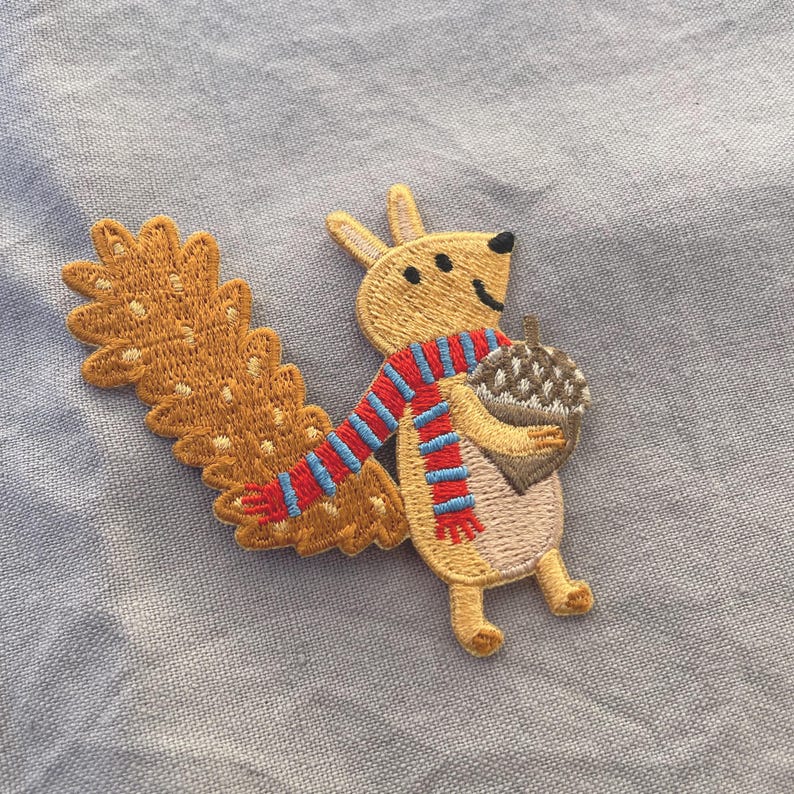 May include: Embroidered patch featuring a cartoon squirrel holding an acorn. The squirrel is tan with a red, white, and blue striped scarf. The patch has a detailed, textured design and is set against a light blue background.