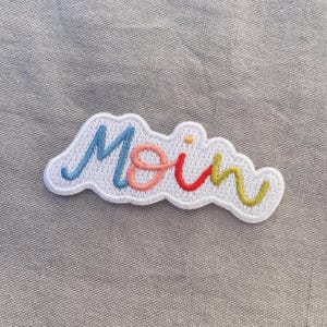May include: Embroidered patch with the word "Moin" in colorful script. The word is in blue, pink, red, and yellow, outlined in white, and has a cloud-like shape.