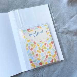 May include: A clear plastic passport holder with a colorful abstract design. The holder has a pocket for a passport and a smaller pocket for other documents. The text "trip pass" is written on the front of the holder.