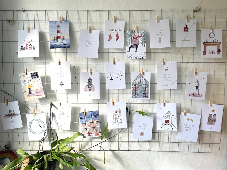 Puede incluir: Una exhibici&oacute;n de tarjetas navide&ntilde;as de acuarela, cada una sujeta con una pinza de madera a una rejilla met&aacute;lica. Las tarjetas presentan ilustraciones festivas, incluyendo un faro, un mu&ntilde;eco de nieve y una chimenea. Una planta en maceta est&aacute; en primer plano.