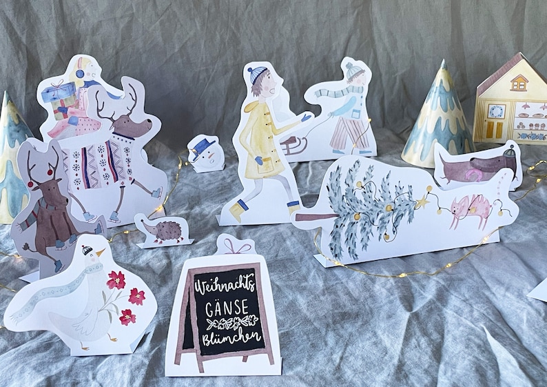May include: A set of paper cutouts featuring a winter scene with a deer, a goose, a hedgehog, a snowman, a Christmas tree, a house, a person pulling a sled, a person carrying gifts, and a sign that reads "Weihnachts G&auml;nse bl&uuml;mchen".