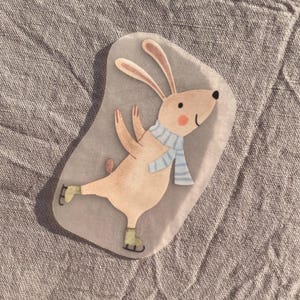 Op de afbeelding: Waterverfillustratie van een lichtbruin konijn dat schaatst. Het konijn draagt een blauw-wit gestreepte sjaal en groene schaatsen. Het konijn heeft roze wangen en een kleine roze staart.