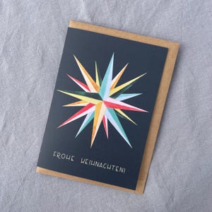 May include: A greeting card with a navy blue background and a colorful starburst design in shades of yellow, red, blue, and green. The card is inside a brown envelope, and the text "FROHE WEIHNACHTEN!" is printed at the bottom.