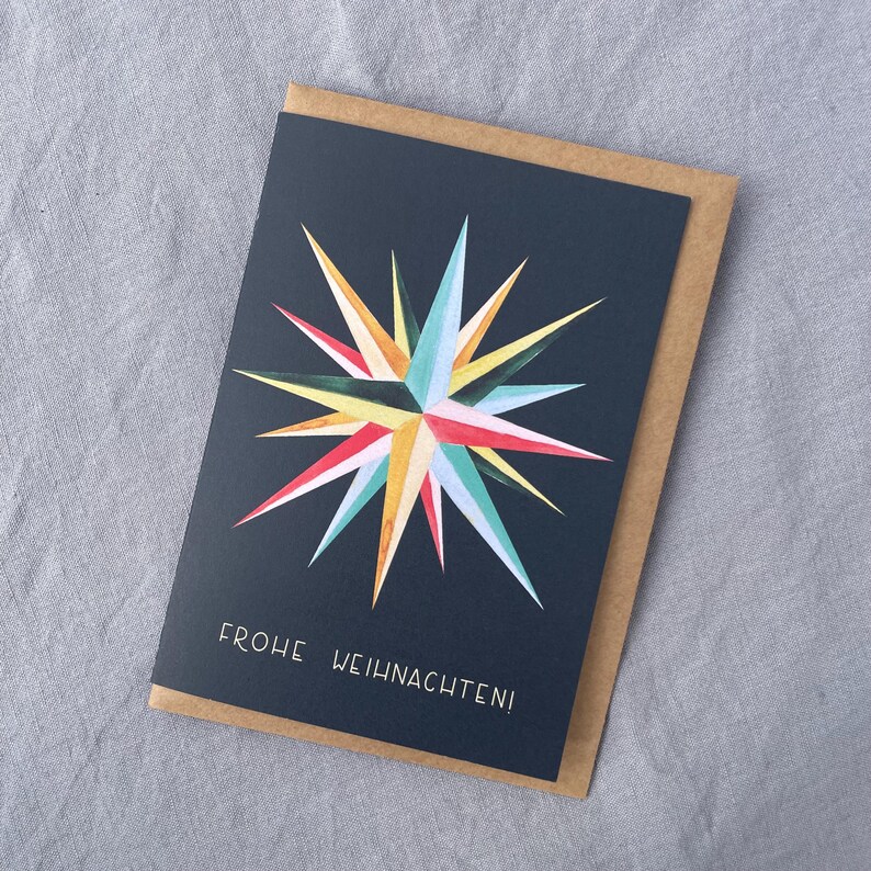 May include: A greeting card with a navy blue background and a colorful starburst design in shades of yellow, red, blue, and green. The card is inside a brown envelope, and the text "FROHE WEIHNACHTEN!" is printed at the bottom.