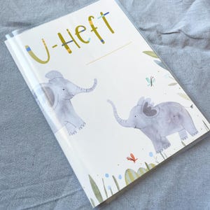 May include: A white binder with a clear plastic cover. The front cover has the text "U-Heft" in yellow letters. There are two watercolour illustrations of elephants on the cover, along with green leaves and blue and red butterflies.