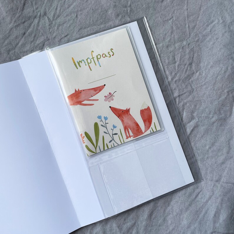 Może przedstawiać: Biała książeczka z przezroczystą plastikową kieszenią, w kt&oacute;rej znajduje się karta z napisem "Impfpass" oraz ilustracjami dw&oacute;ch lis&oacute;w, kwiat&oacute;w i liści. Lisy są pomarańczowe z białymi akcentami.