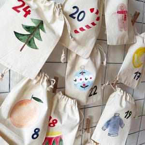 May include: A collection of off-white cotton drawstring bags featuring watercolor illustrations and numbers. Designs include a green tree with 24, a candy cane with 20, a gingerbread person, a blue ornament with 22, a peach with 8, and a sweater with 19.