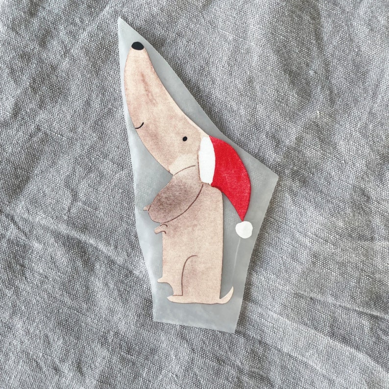 May include: A brown and white cartoon dachshund wearing a red Santa hat with white trim. The image is on a gray fabric background.