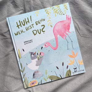 Könnte beinhalten: Ein Kinderbuch mit festem Einband mit dem Titel Huh! Wer bist denn du? von Hannah Siehoff. Das Cover zeigt eine Aquarellillustration eines rosa Flamingos und eines kleinen grauen Vogels, der eine Blume in einem blauen Teich hält.