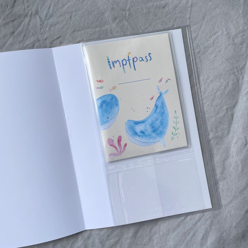 Może przedstawiać: Przezroczysty plastikowy folder z białą kartką w środku. Na kartce znajduje się akwarelowa ilustracja dw&oacute;ch niebieskich wieloryb&oacute;w i napis "Imptpass".