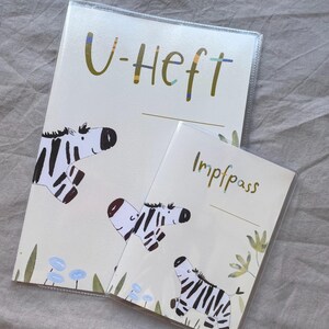 May include: Two protective covers for documents, one larger labelled U-Heft and one smaller labelled Impfpass. Both feature a cream background with illustrations of zebras, green foliage, and small blue flowers. The text is in a dark green font.
