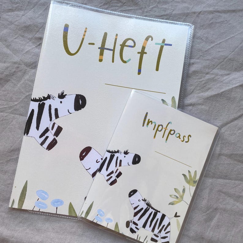 May include: Two protective covers for documents, one larger labelled U-Heft and one smaller labelled Impfpass. Both feature a cream background with illustrations of zebras, green foliage, and small blue flowers. The text is in a dark green font.