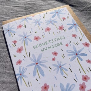 May include: A greeting card with a floral design. The card features light blue flowers with red centers and small pink flowers scattered across a white background. The text "GEBURTSTAGS W&Uuml;NSCHE" is printed in green.