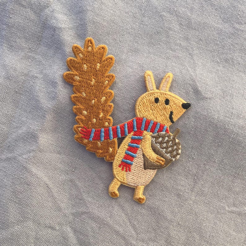 May include: Embroidered patch featuring a cartoon squirrel with a fluffy tail and a striped scarf. The squirrel is holding an acorn. The patch is in shades of tan, brown, red, and blue.