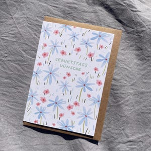 May include: A greeting card with a floral design. The card features light blue flowers and small pink flowers on a white background. The text "GEBURTSTAGS W&Uuml;NSCHE" is printed in green. The card is inside a brown envelope.