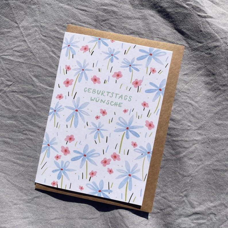 May include: A greeting card with a floral design. The card features light blue flowers and small pink flowers on a white background. The text "GEBURTSTAGS W&Uuml;NSCHE" is printed in green. The card is inside a brown envelope.