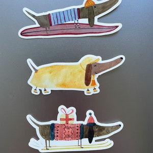 May include: Three dachshund dog stickers with different winter holiday themes. The first sticker shows a dachshund wearing a blue and white striped shirt and a yellow hat, riding a surfboard. The second sticker shows a dachshund wearing a yellow raincoat and a yellow hat, with a bell around its neck. The third sticker shows a dachshund wearing a red and white sweater and a red hat, carrying a gift and skiing on skis.