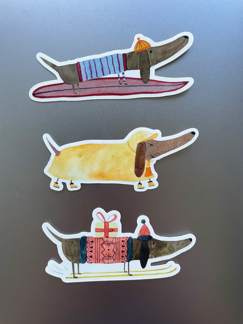 May include: Three dachshund dog stickers with different winter holiday themes. The first sticker shows a dachshund wearing a blue and white striped shirt and a yellow hat, riding a surfboard. The second sticker shows a dachshund wearing a yellow raincoat and a yellow hat, with a bell around its neck. The third sticker shows a dachshund wearing a red and white sweater and a red hat, carrying a gift and skiing on skis.