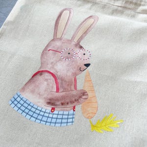 May include: A brown bunny wearing red suspenders and blue and white checkered pants holds a carrot in its paws. The bunny is wearing round glasses with red and white frames. The bunny is on a beige canvas tote bag.