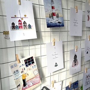 Puede incluir: Una exhibici&oacute;n enrejada de impresiones art&iacute;sticas en acuarela, cada una sujeta con una pinza de madera. Las impresiones presentan ilustraciones de casas, un faro y una persona horneando. Algunas impresiones tienen texto y n&uacute;meros.