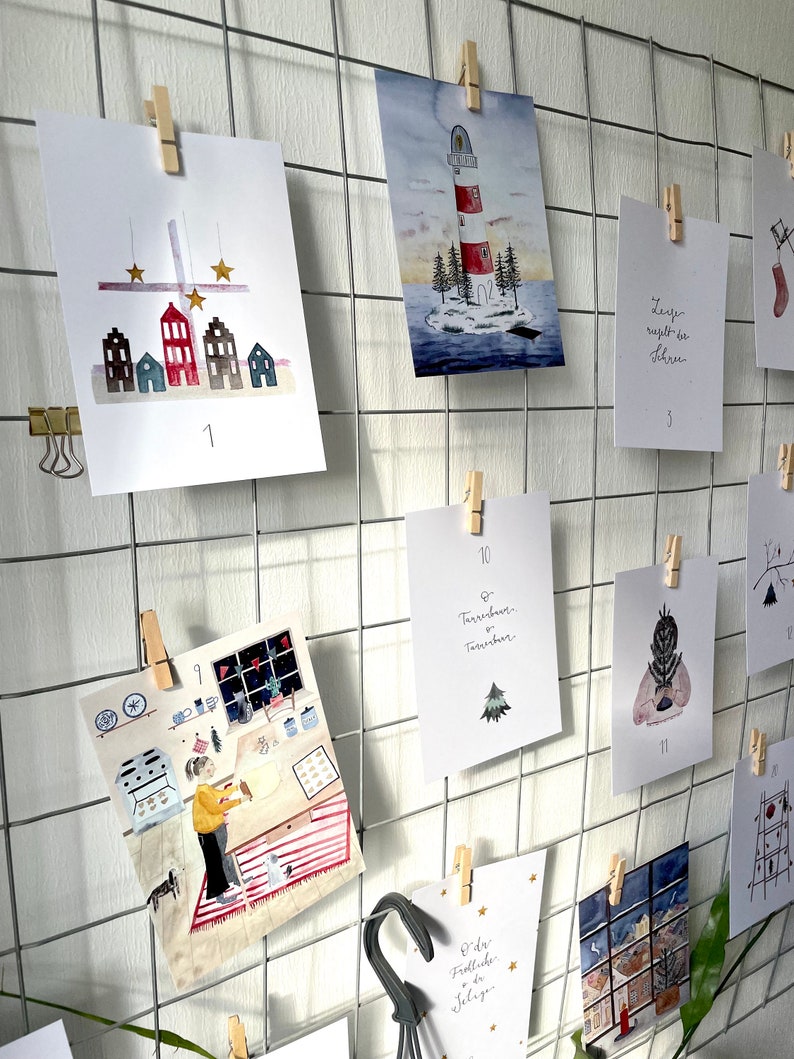 Puede incluir: Una exhibici&oacute;n enrejada de impresiones art&iacute;sticas en acuarela, cada una sujeta con una pinza de madera. Las impresiones presentan ilustraciones de casas, un faro y una persona horneando. Algunas impresiones tienen texto y n&uacute;meros.