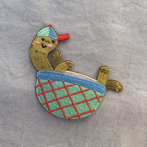 May include: Embroidered patch of a cartoon pickle in a blue and red baseball cap, lounging in a green and red basket. The pickle has a smiling face and yellow feet. The patch is on a light blue fabric.