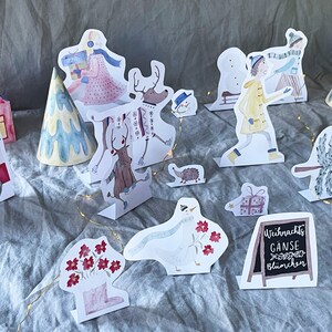 May include: A set of Christmas-themed paper cutouts, including a reindeer, a snowman, a goose, a tree, a house, and a sign that reads "Weihnachts G&auml;nse Bl&uuml;mchen".