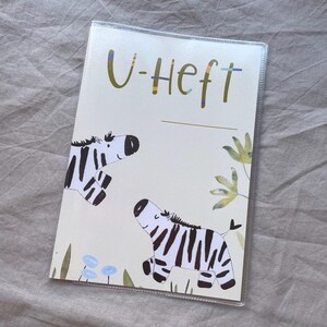 May include: A rectangular protective cover for a medical booklet featuring the text U-Heft in olive green. The design includes two illustrated zebras with black and white stripes, small blue flowers, and green foliage on a cream-coloured background.