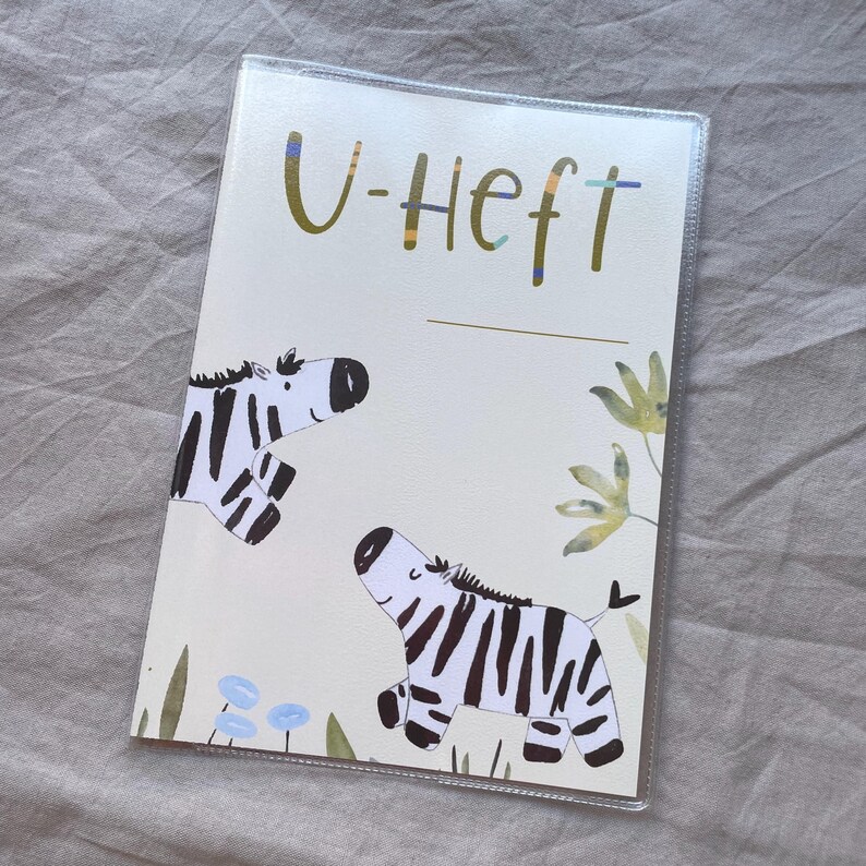 May include: A rectangular protective cover for a medical booklet featuring the text U-Heft in olive green. The design includes two illustrated zebras with black and white stripes, small blue flowers, and green foliage on a cream-coloured background.