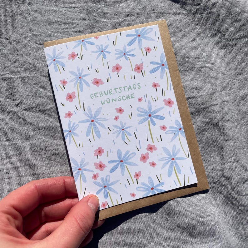 May include: A birthday greeting card with a floral design. The card features light blue flowers and pink flowers on a white background. The words "GEBURTSTAGS W&Uuml;NSCHE" are printed in green. The card is held in a person's hand.