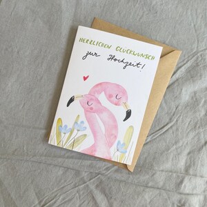 May include: A wedding greeting card featuring a watercolour illustration of two pink flamingos with black beaks. The card has the text "HERZLICHEN GL&Uuml;CKWUNSCH zur Hochzeit!" in green and black. A small red heart is above the flamingos. The card is inside a brown envelope.
