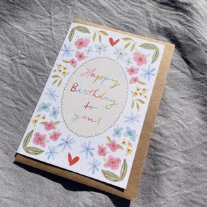 Może przedstawiać: Kartka urodzinowa z kwiatowym wzorem. Karta ma białe tło z kolorową kwiatową obw&oacute;dką i słowami "Happy Birthday to you!" w tęczowej czcionce. Karta jest umieszczona na brązowym tle papieru.