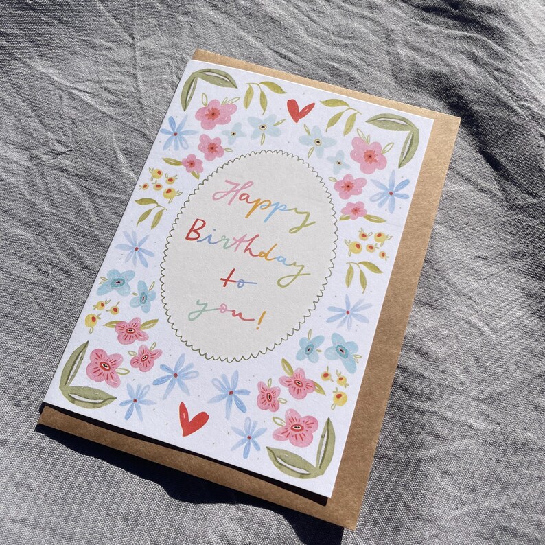 Może przedstawiać: Kartka urodzinowa z kwiatowym wzorem. Karta ma białe tło z kolorową kwiatową obw&oacute;dką i słowami "Happy Birthday to you!" w tęczowej czcionce. Karta jest umieszczona na brązowym tle papieru.