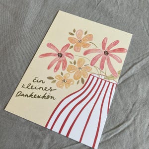 May include: A greeting card featuring a watercolour illustration of flowers in a vase. The vase has red and white stripes. The flowers are pink and orange. The card includes the text "Ein kleines Dankesch&ouml;n".